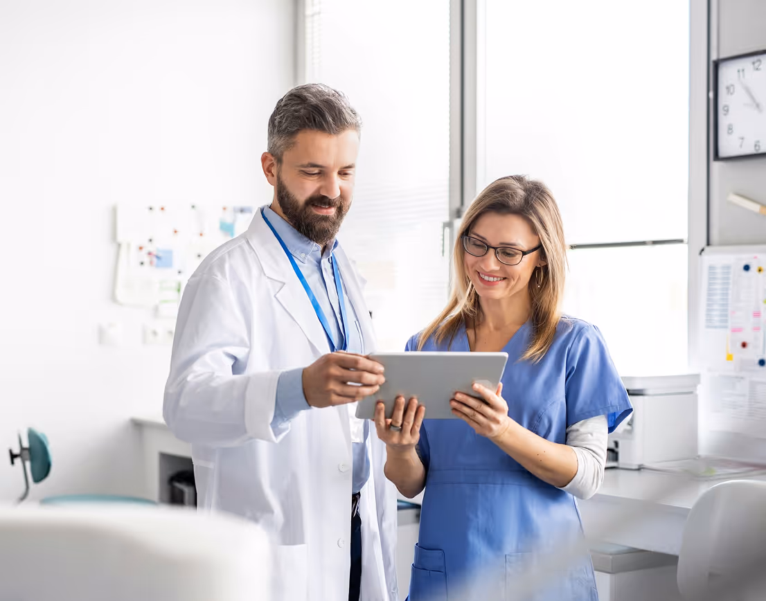 Male doctor and female nurse smiling while looking at a tablet in a bright medical office.