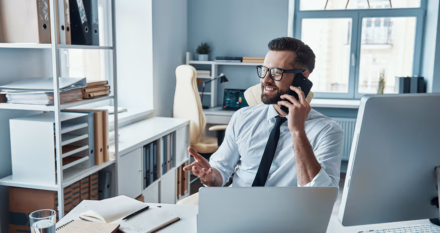 Smiling businessman in glasses talking on phone at desk with laptop and open notebooks in office.