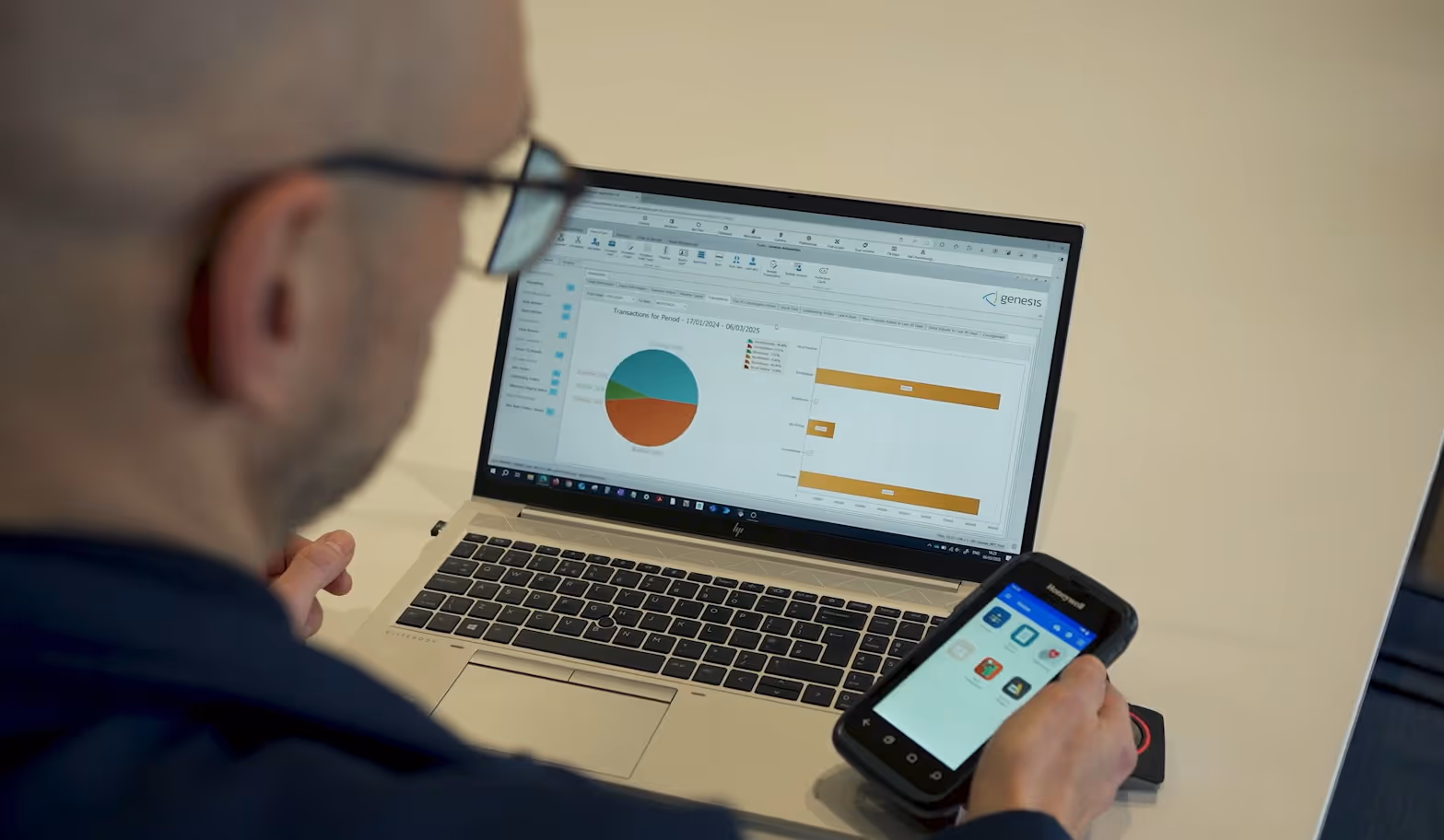 Man using a handheld device while viewing financial charts on a laptop screen with the Genesis logo.