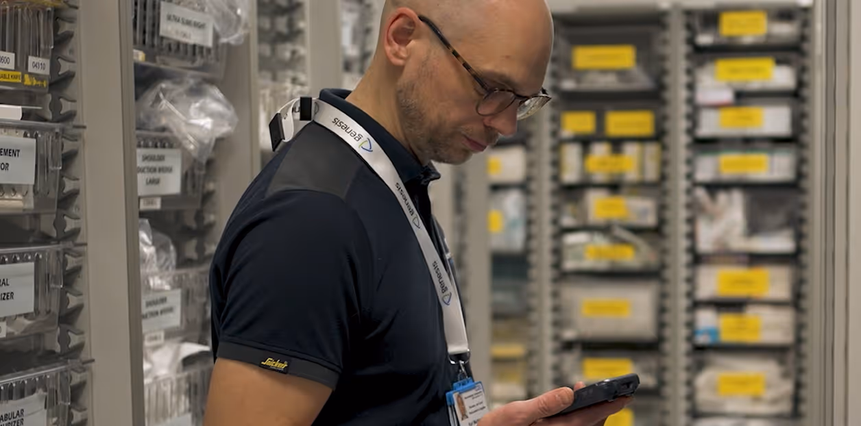 Man with glasses and lanyard checking a smartphone in a storage room with labeled shelves.