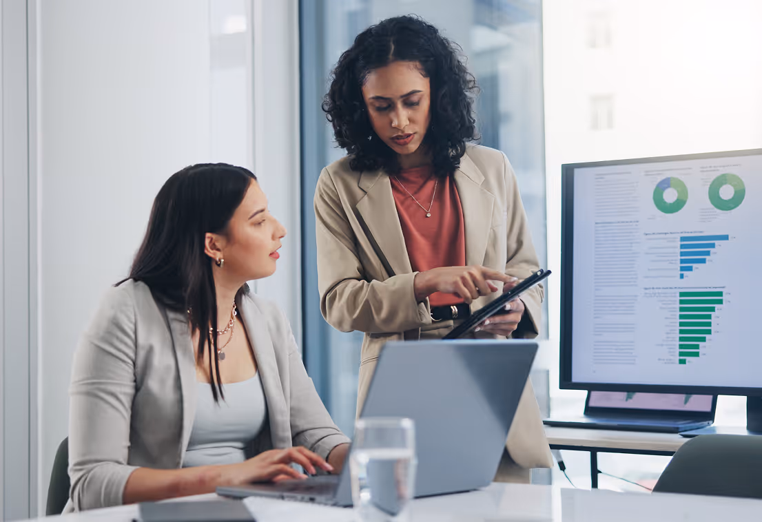 Two businesswomen discussing data on a tablet by a screen showing charts and graphs in an office.