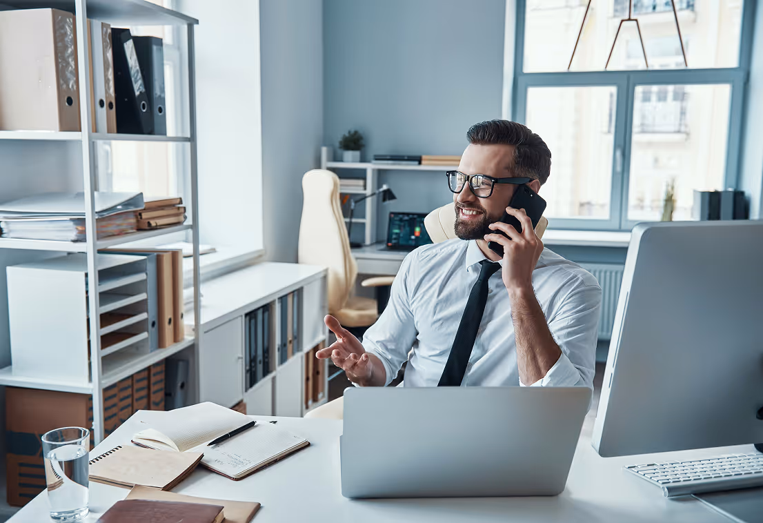 Smiling businessman in glasses talking on phone at desk with laptop and office supplies.