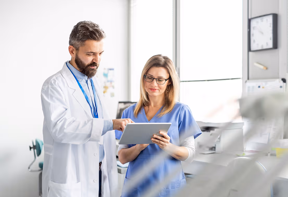 Male doctor in white coat showing a tablet to a female nurse in blue scrubs in a medical office.