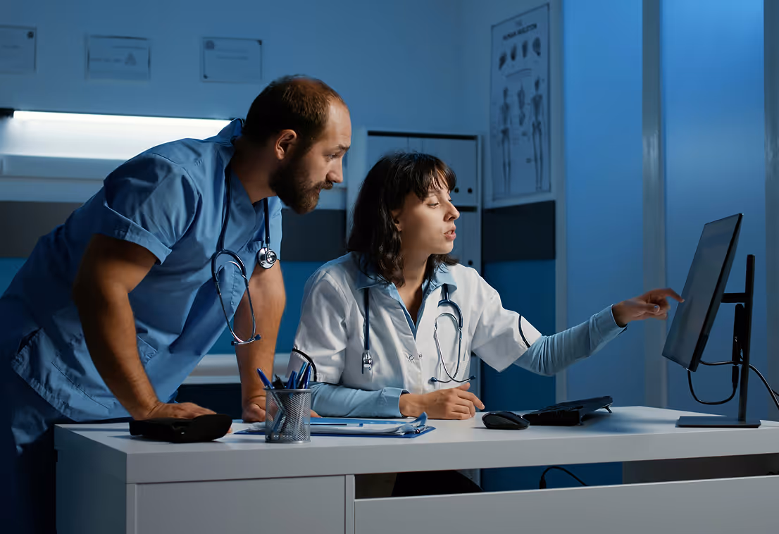 Two doctors in scrubs discussing patient data on a computer in a medical office.