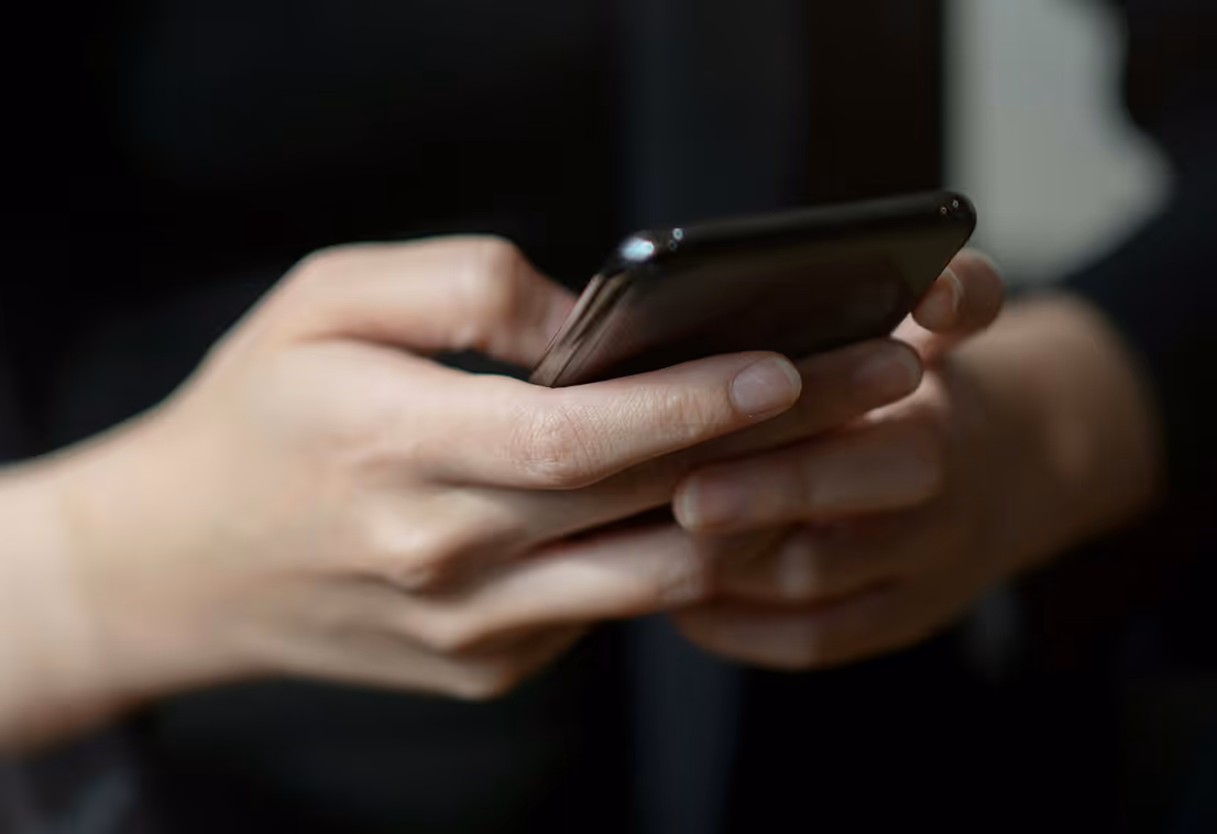 Close-up of both hands holding and using a black smartphone with a dark background.