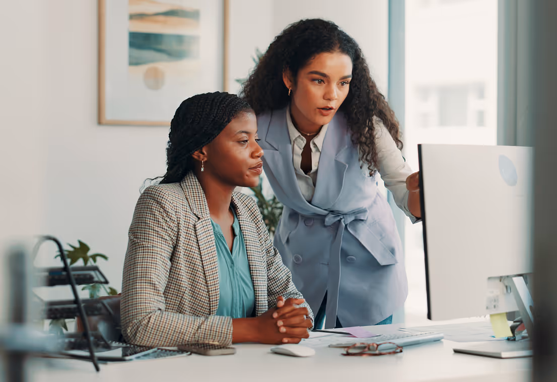 Two women work together looking at a computer screen in a bright office setting.