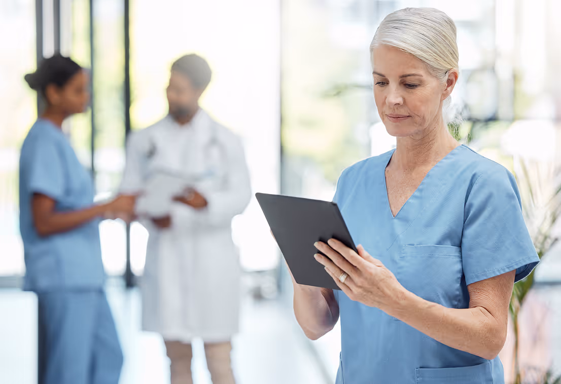 Female nurse in blue scrubs using a tablet with a doctor and nurse talking blurred in background.