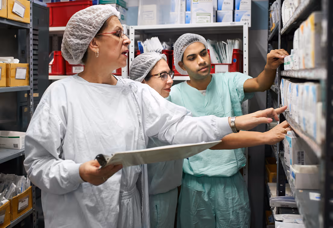Three healthcare professionals in protective clothing reviewing medical supplies on shelves in a storage room.