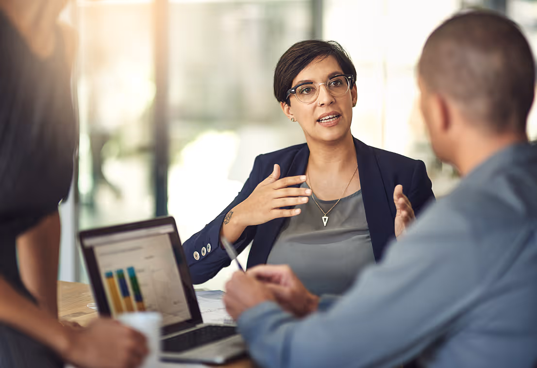 Woman explaining data on a laptop to two colleagues in an office meeting.