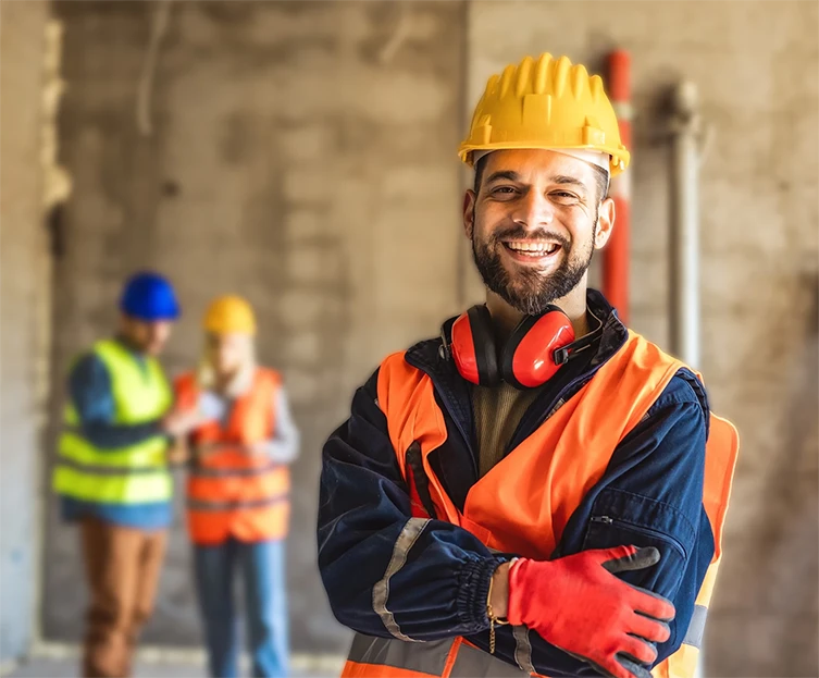 Operaio edile sorridente con casco giallo, giubbotto arancione e protezioni acustiche appuntate al collo in un cantiere.