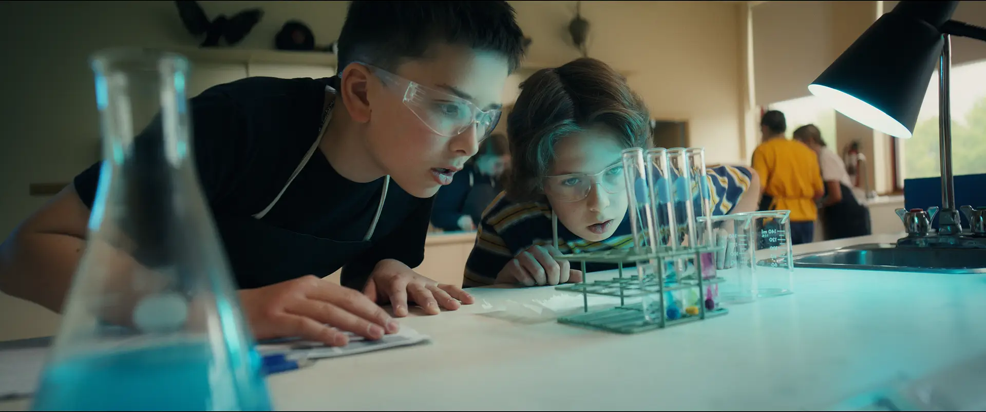 Deux élèves à lunettes de sécurité observant des éprouvettes colorées en laboratoire — publicité Collège Mont-Sacré-Cœur