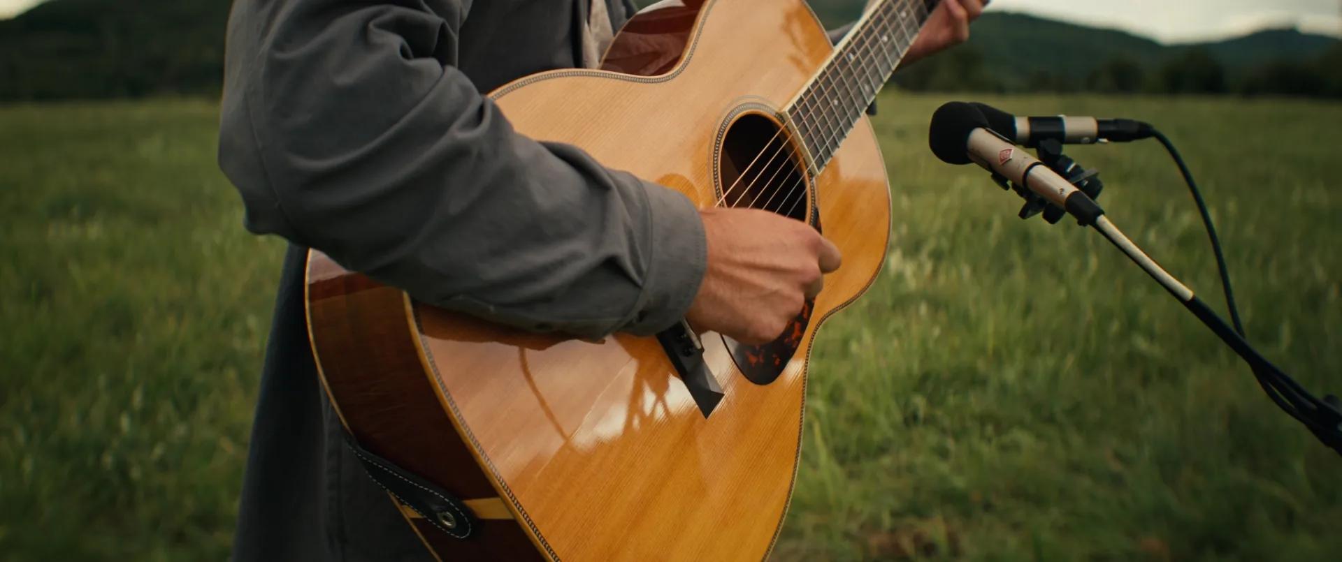 Main pinçant les cordes d'une guitare acoustique devant un micro dressé dans l'herbe, tournage de vidéoclip