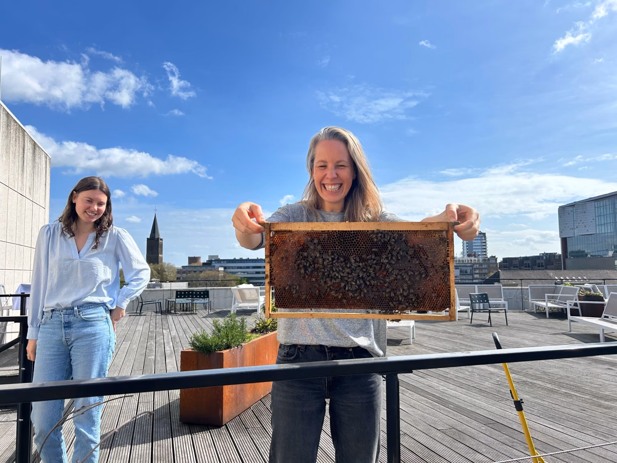 A smiling beekeeper holds up a honeycomb frame covered in bees on a building rooftop while a tenant looks on, with a city skyline behind.