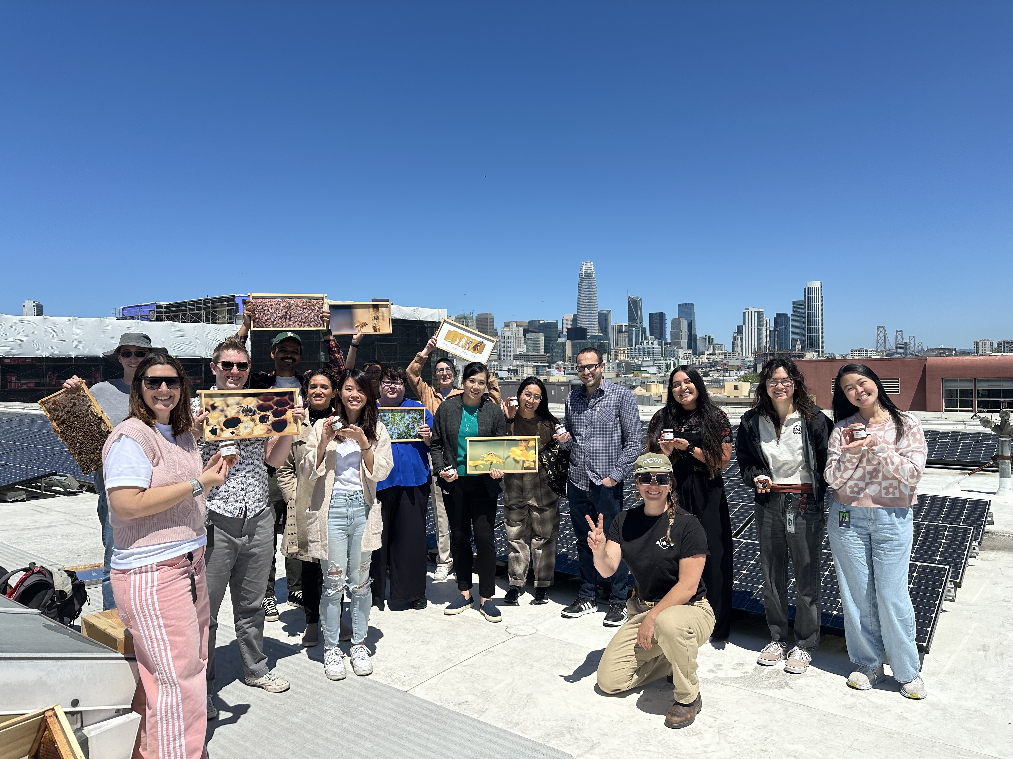 Group of tenants holding honeycomb frames and honey jars at an Alvéole rooftop workshop in San Francisco, with the downtown skyline in the background