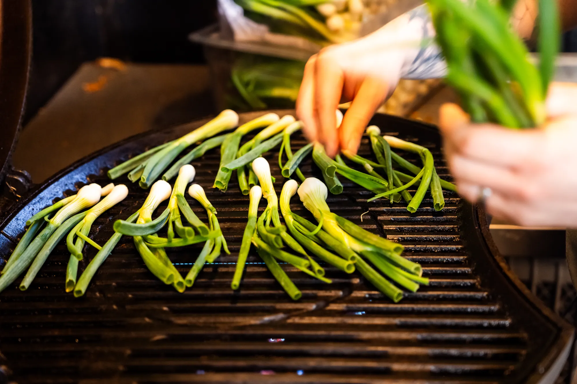 Hænder lægger grønne forårsløg på en varm grillrist til madlavning.
