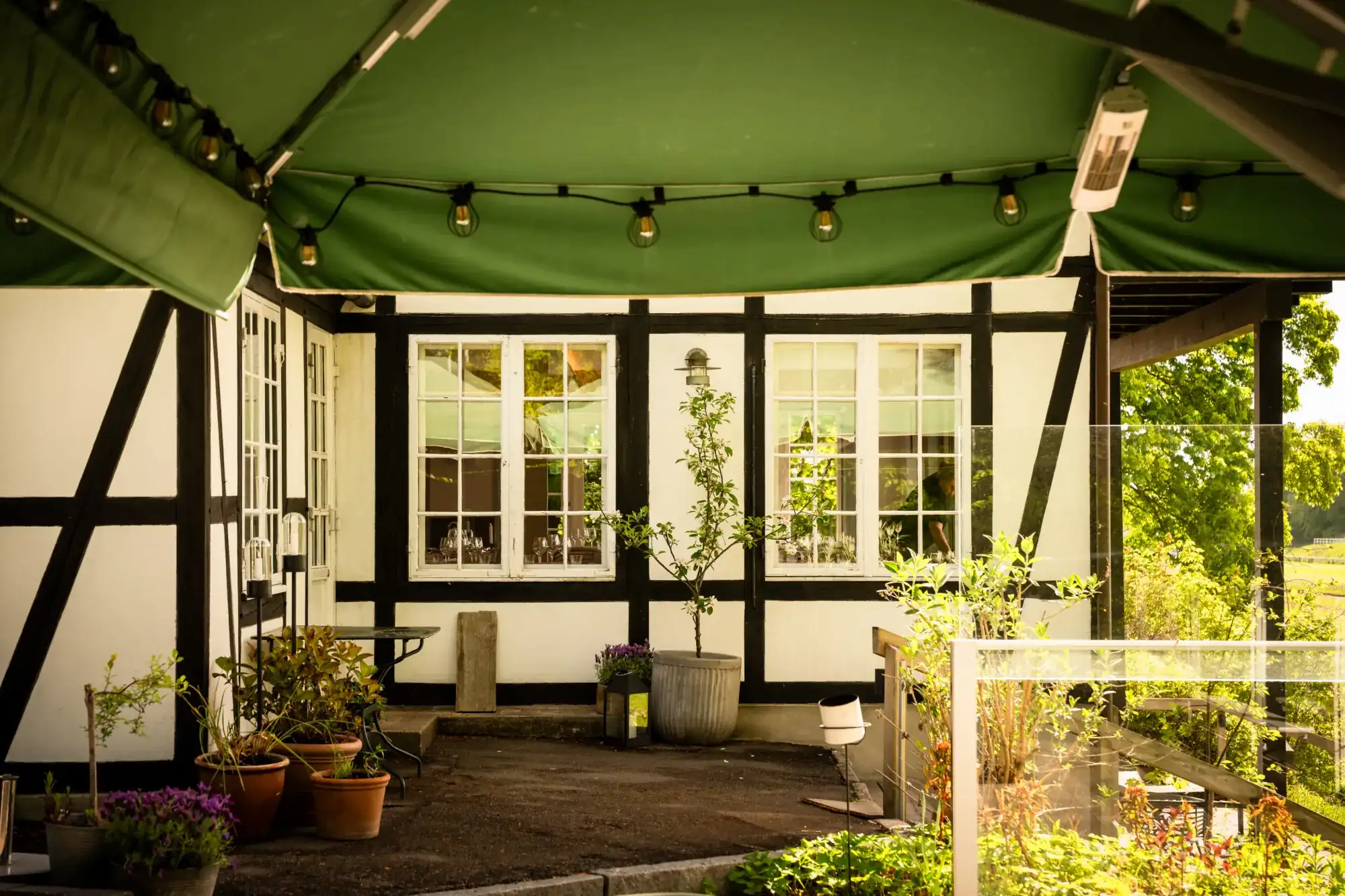 Terrasse med planter under grøn markise ved bindingsværkshus med store vinduer.