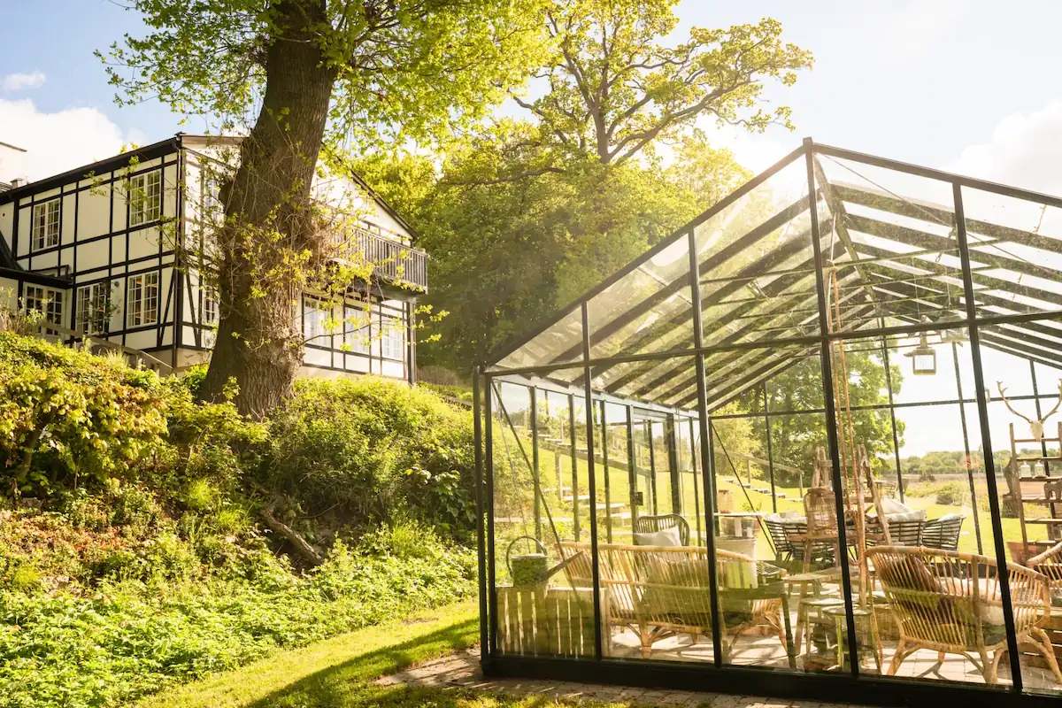 Glashus med møbler i en solrig have nær et hvidt hus med sorte trærammer.