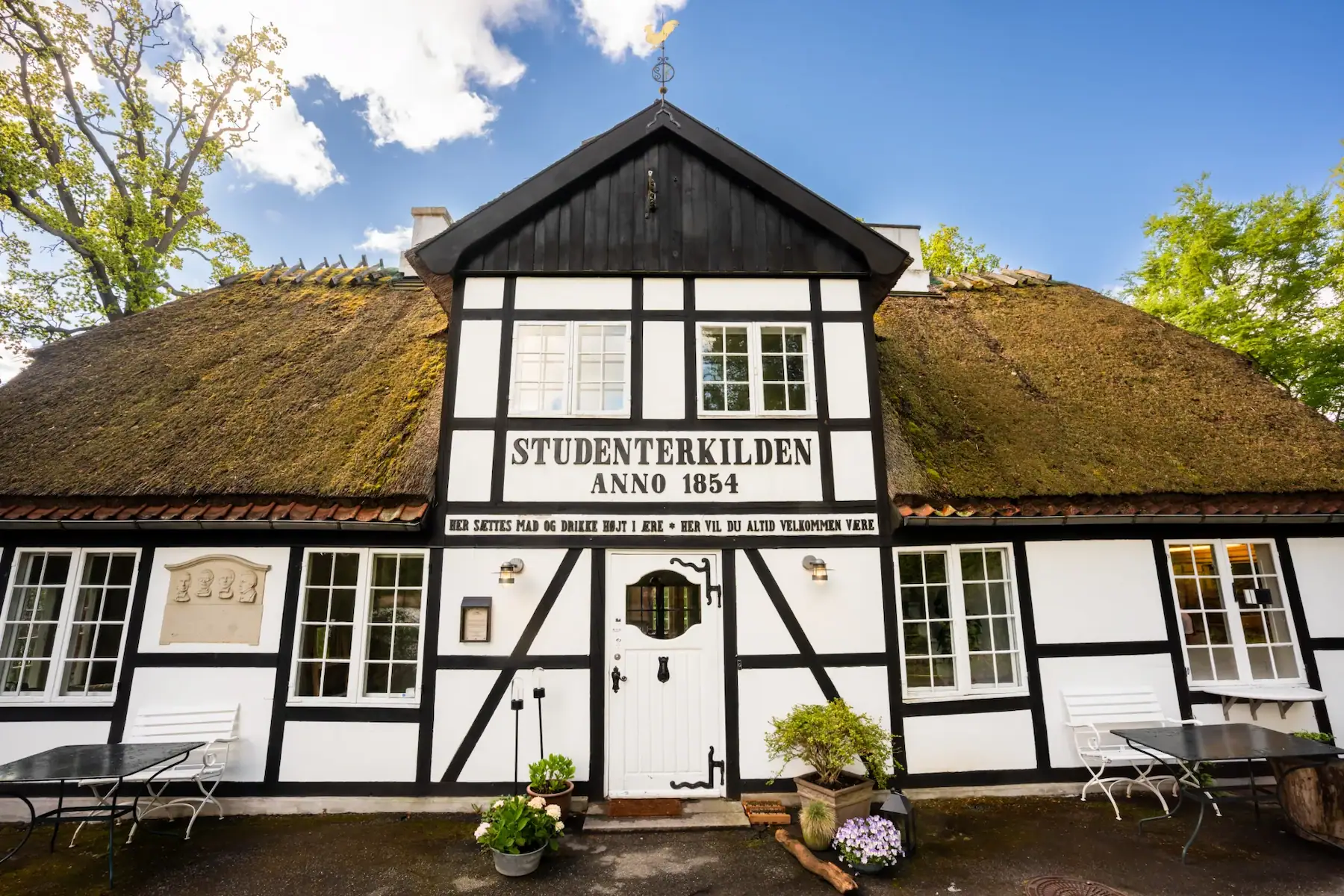 Traditionelt bindingsværkshus med mosstag og skilt 'STUDENTERKILDEN ANNO 1854' på facaden.