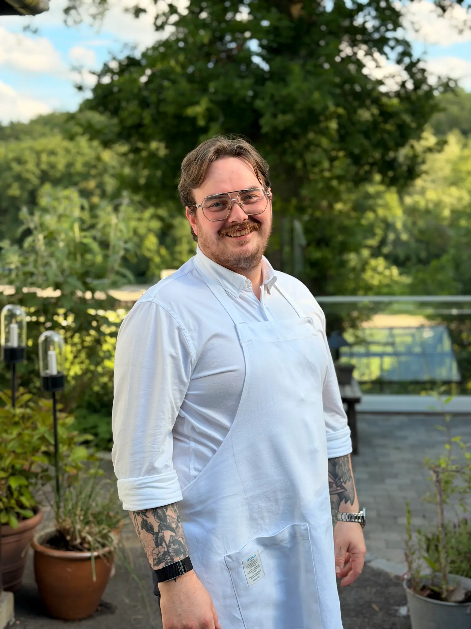Mand med hvid skjorte, forklæde og tatoverede arme smilende udenfor i haven.
