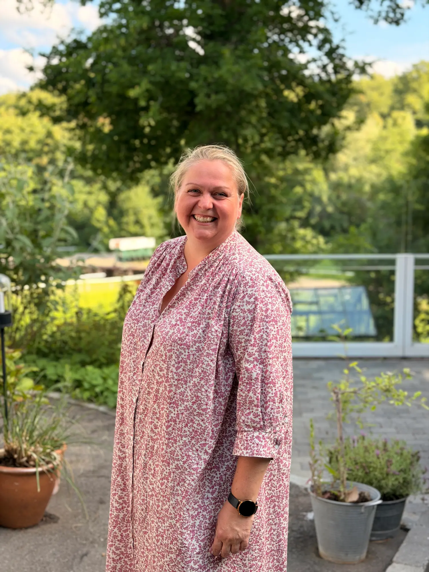 Smilende kvinde i lyserød blomstret kjole udendørs med grøn baggrund og potteplanter.