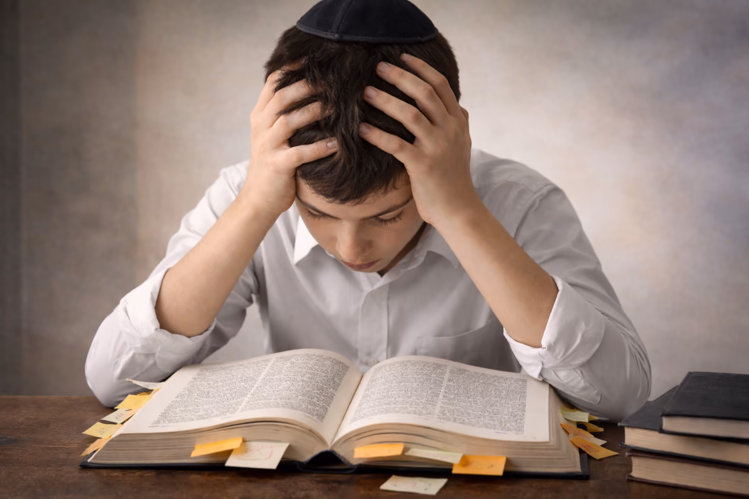 A young teen boy is bent over a Torah book marked with several sticky notes, head between his hands