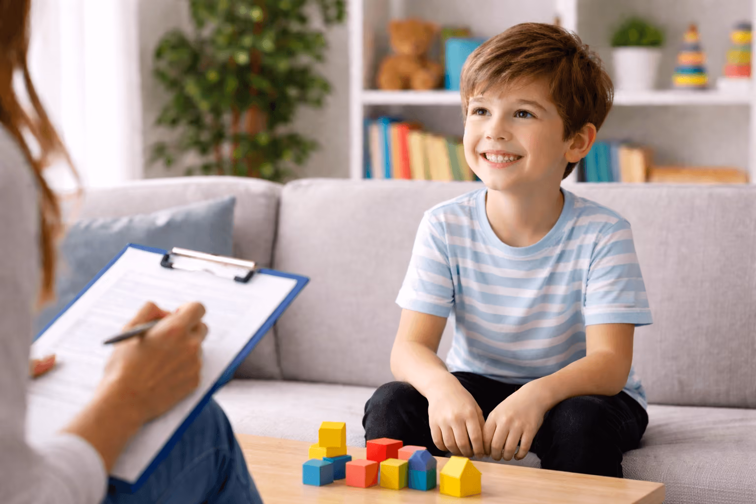 A smiling young child sits on a couch playing with colorful blocks while a clinician takes notes on a clipboard, illustrating a child developmental evaluation in a supportive, home-like setting