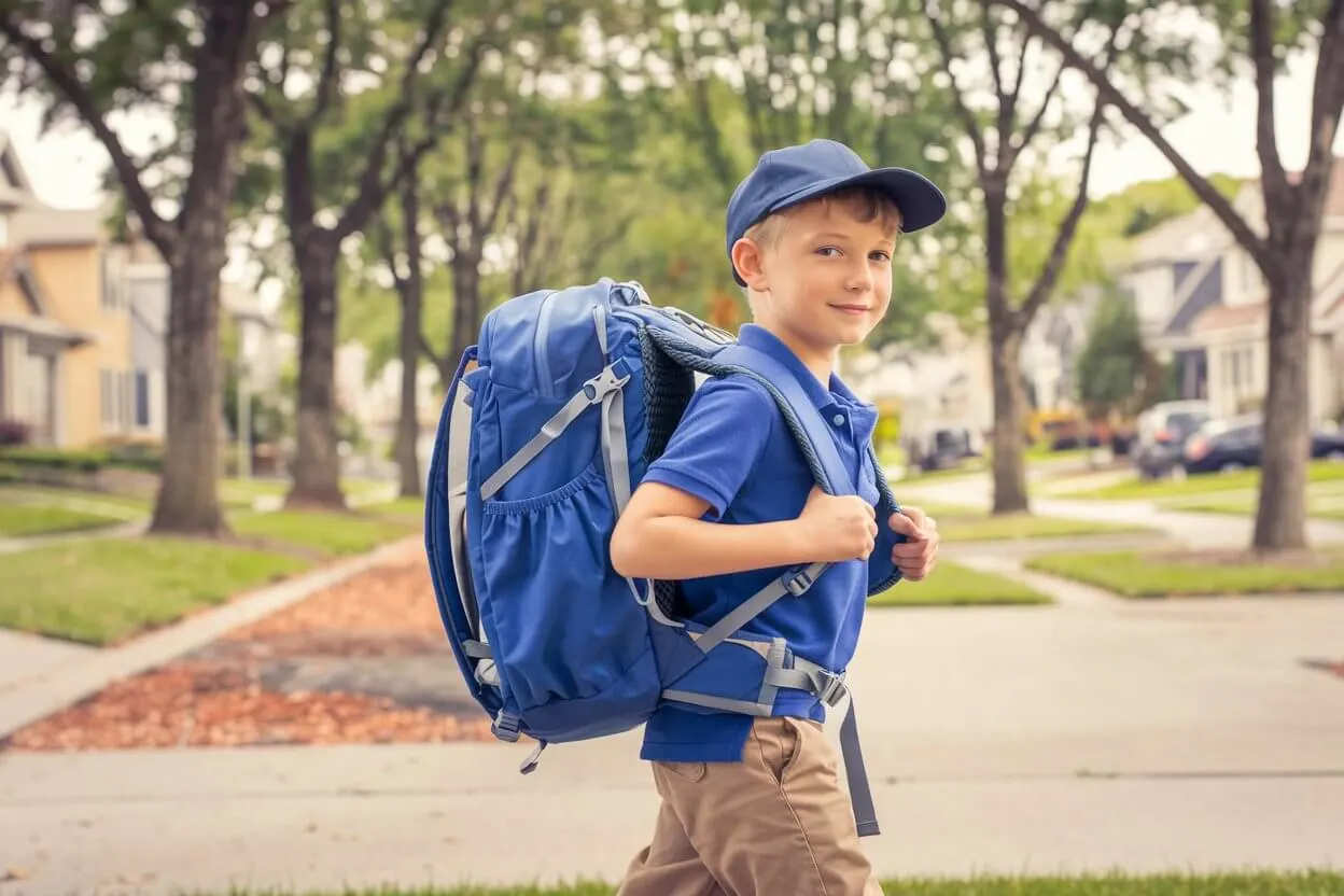 A young boy bravely holds his backpack with a small smile, as he heads off for his first day of school.