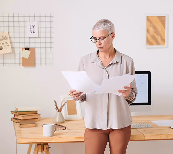 Person with short gray hair and glasses reading papers in a tidy home office with desk and computer.