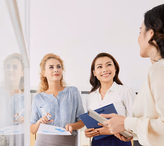 Three professional women discussing work and smiling in a bright office setting.
