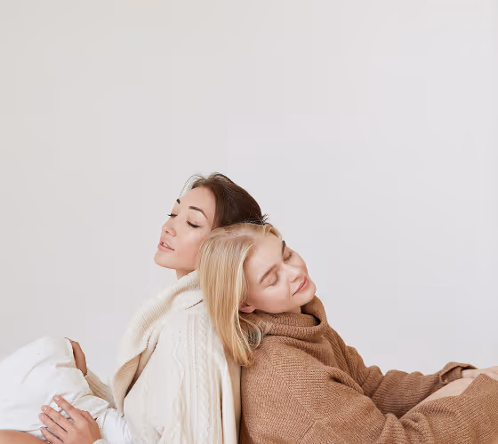 Two women sitting back to back with eyes closed, wearing cozy sweaters against a plain background.