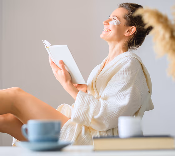 Smiling woman in a bathrobe with eye patches reads a book while sitting relaxed indoors.