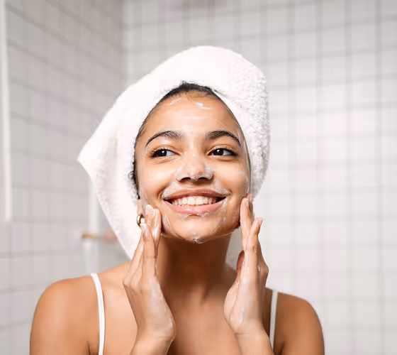 Smiling woman with a white towel wrapped on her head applying facial cleanser foam in a tiled bathroom.