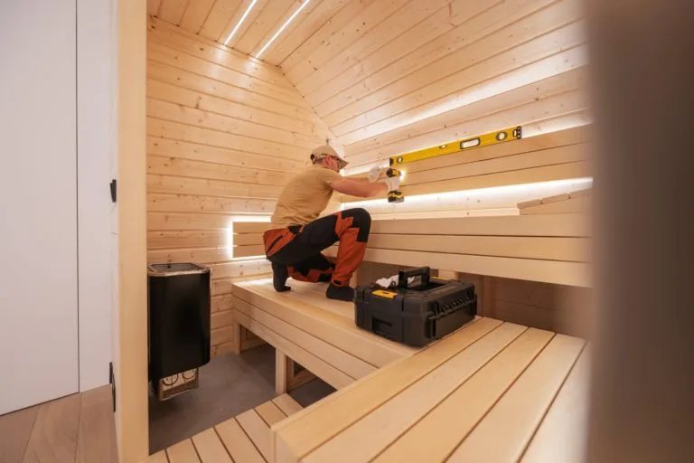 A man using a level tool while working inside a modern wooden sauna with LED lighting and tool box.