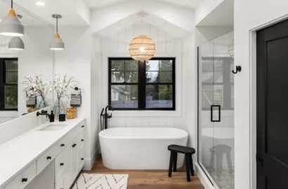 Modern bathroom with white freestanding tub, black stool, glass shower, and double vanity with pendant lights.