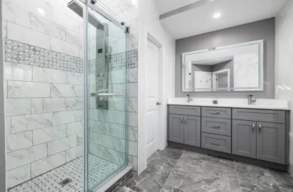 Modern bathroom with glass shower enclosure, marble tile walls, and gray vanity with double sinks and large mirror.
