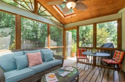 Sunroom with wooden ceiling, large windows, blue sofa, round table, and two cushioned chairs.