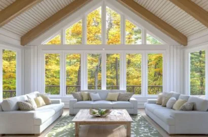 Bright sunroom with large windows, wooden beams, three white sofas, and a wooden coffee table.