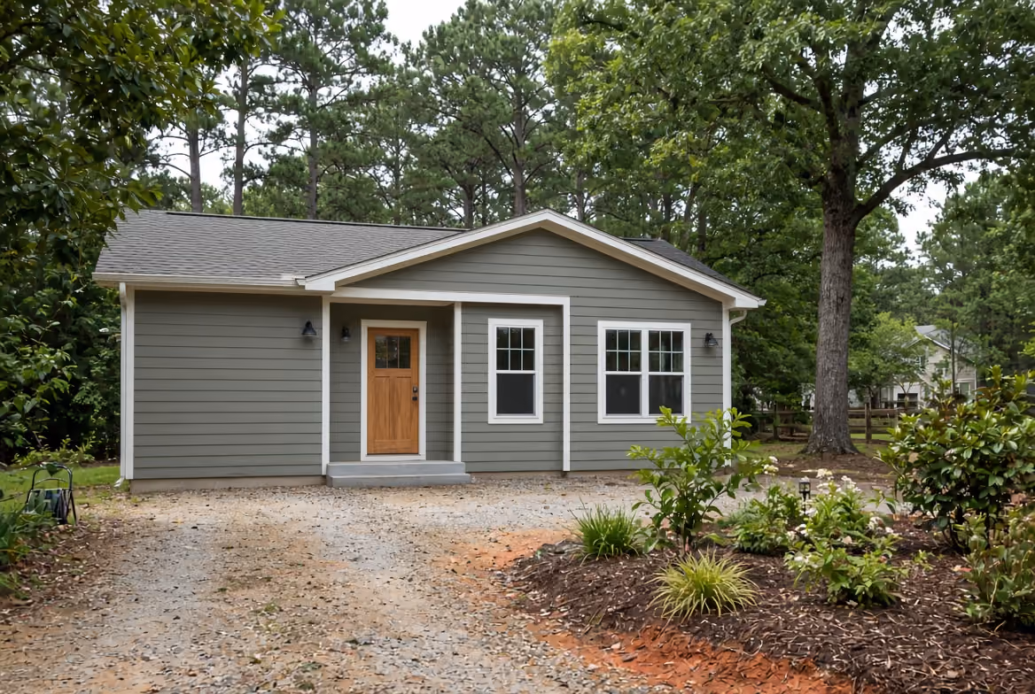 Covered porch with decorative white columns, gray decking, and beige siding beside green trees.