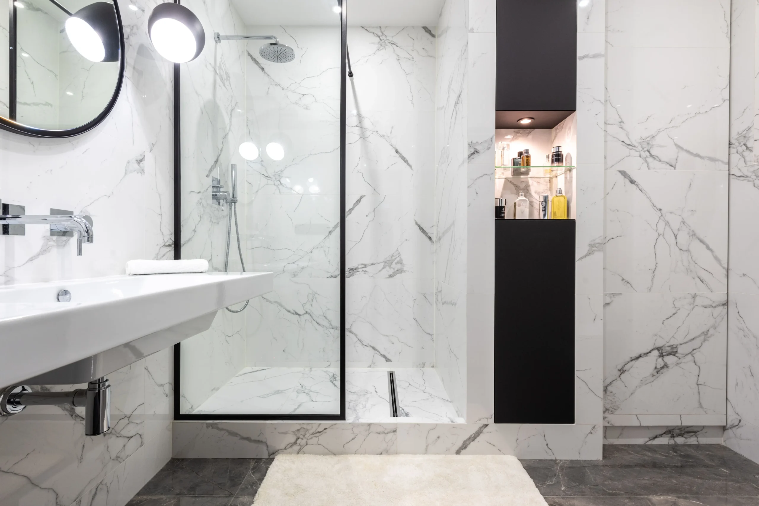 Modern bathroom with marble walls, walk-in glass shower, white sink, and black shelving with toiletries.