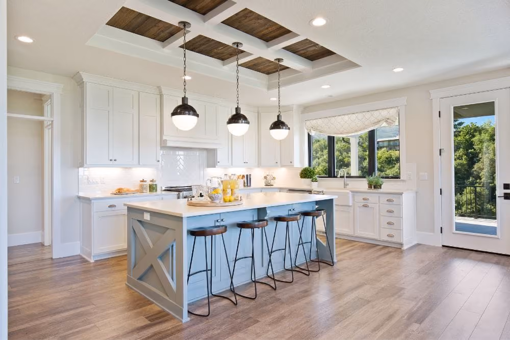 Bright farmhouse kitchen with white cabinets, blue island, wooden stools, and pendant lights on wooden ceiling panels.