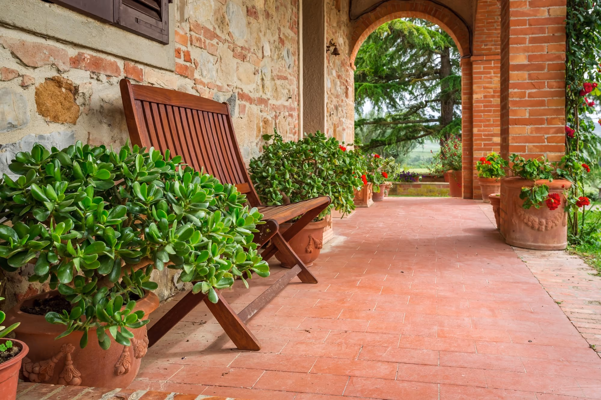 Wooden bench on a brick porch with green plants in pots and an arched stone wall.