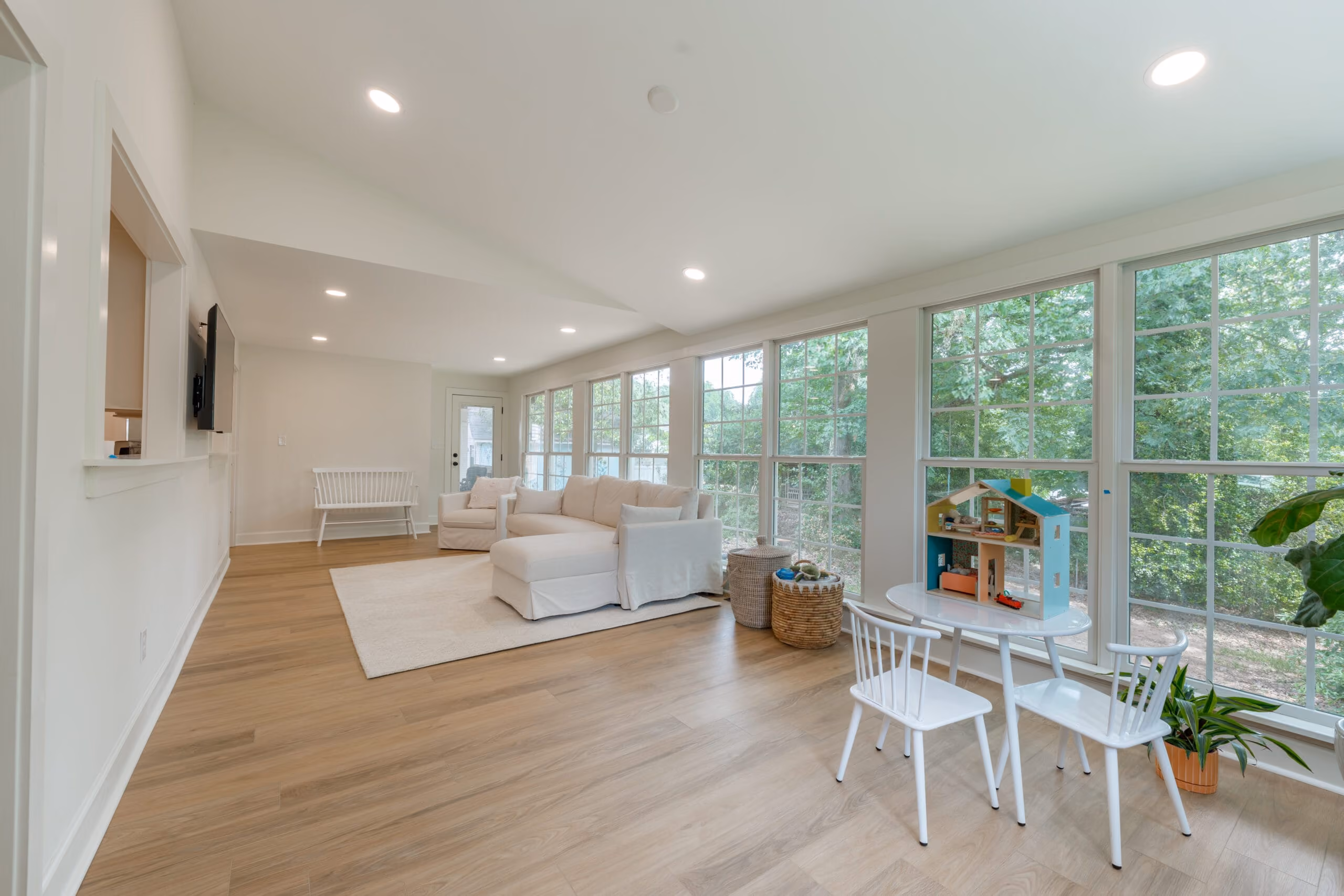 Bright living room with large windows, white sofa, armchair, small table with chairs and dollhouse.