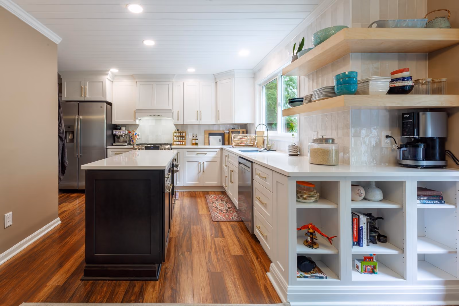 Modern kitchen with white cabinetry, black island with white countertop, stainless steel refrigerator, dishwasher, wooden floors, open shelves holding dishes, and built-in cubbies with books and decorative items.