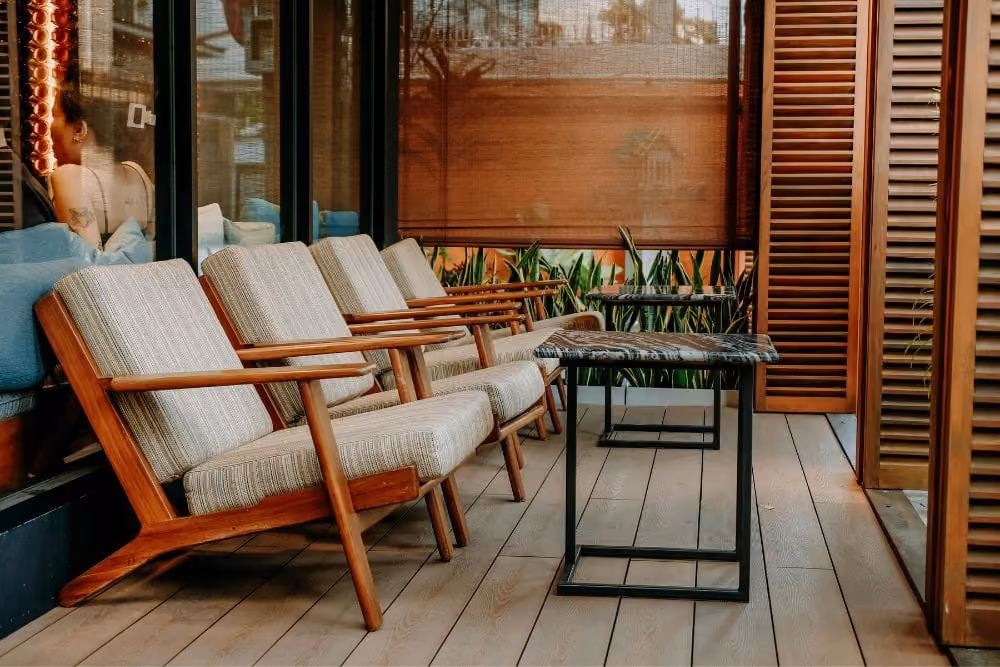 Row of wooden armchairs with gray cushions facing two small black tables on a wooden porch.