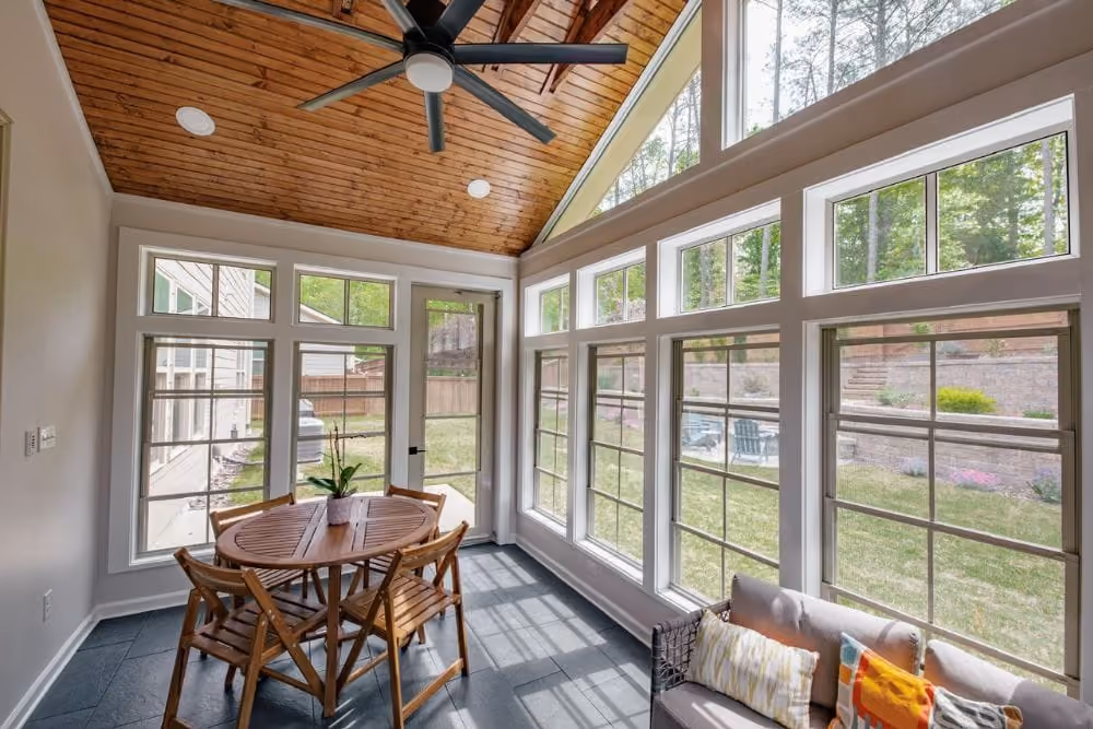 Sunroom with large windows, wooden ceiling fan, round wooden table, chairs, and a cushioned sofa.