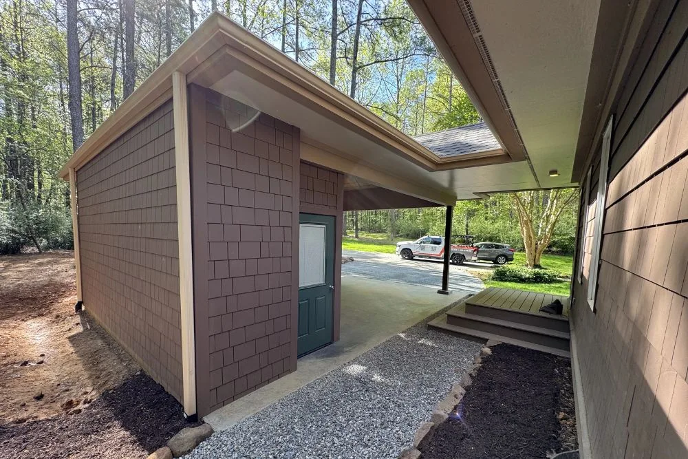 Brown siding storage shed with green door, gravel path, and part of adjacent house with steps.