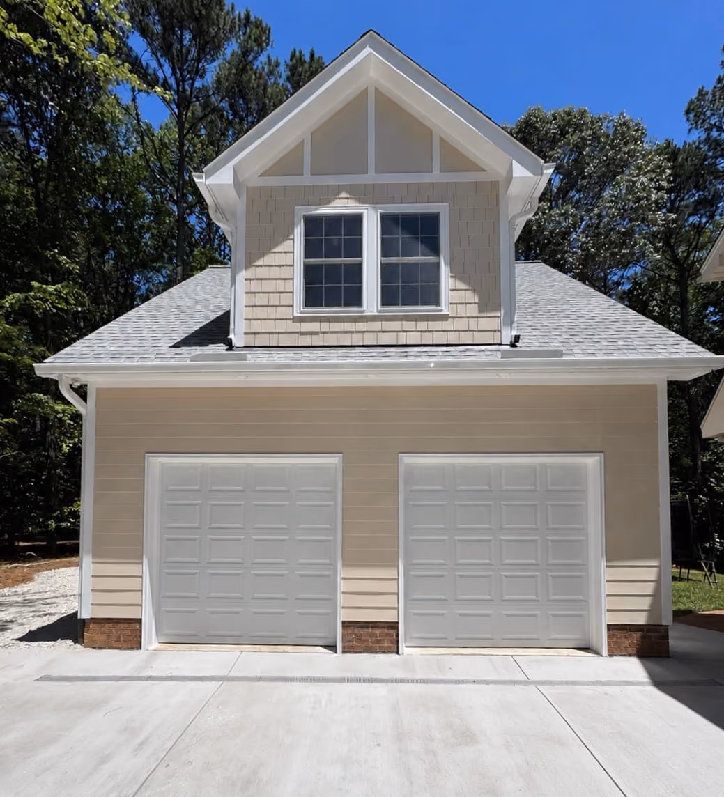 Two-car garage with beige siding and white framed windows above, surrounded by trees under a clear blue sky.