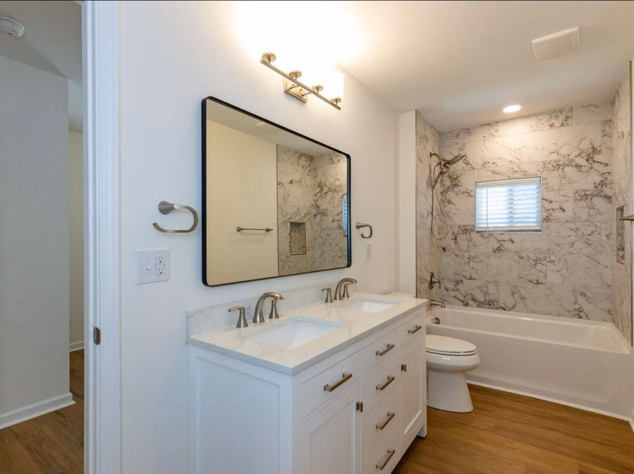 Modern bathroom with double sink white vanity, large rectangular mirror, bathtub with marble tiled walls, toilet, and wooden floor.