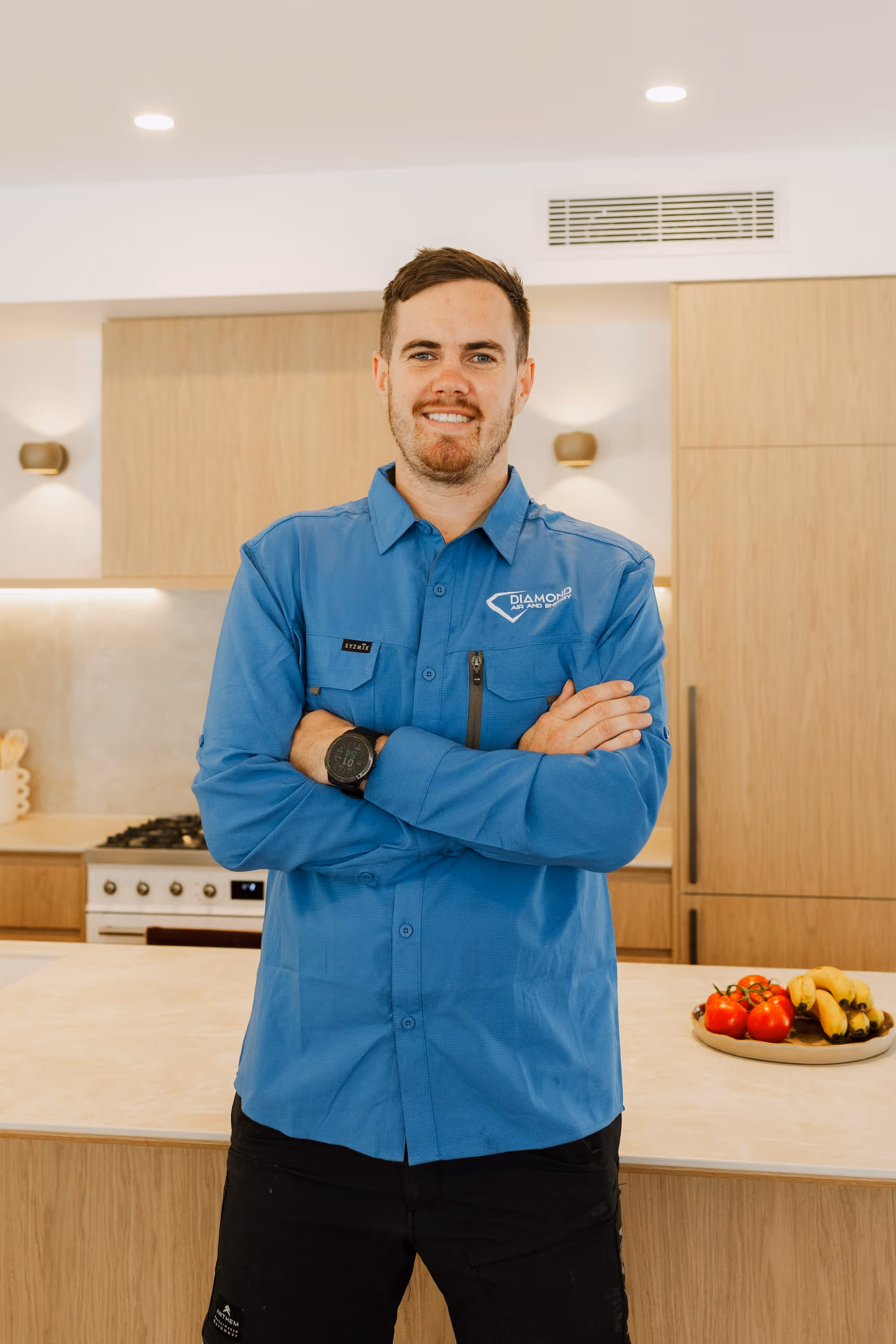 Smiling man in blue Diamond Air and Energy shirt stands with arms crossed in modern kitchen.