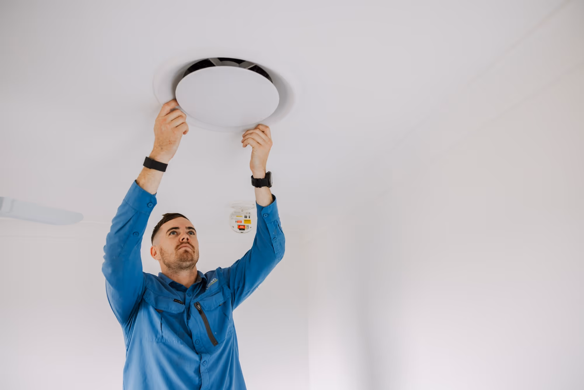 A man in a blue shirt installs or inspects a round ceiling vent or light fixture.